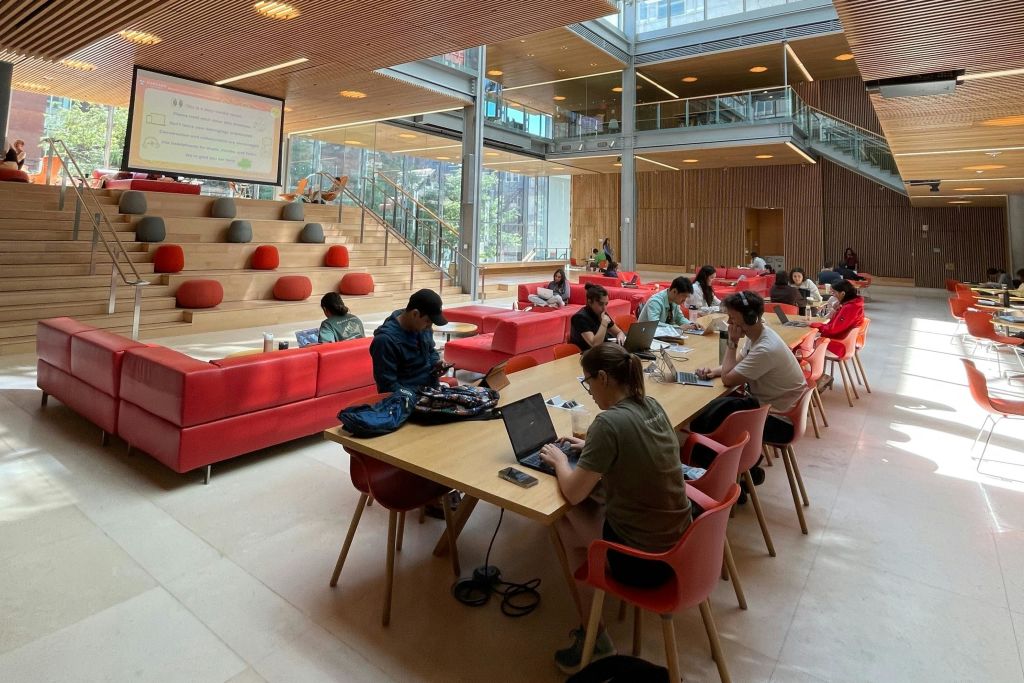 빨간색 소파, 나무 악센트, 커다란 창문이 있는 현대적이고 탁 트인 공간에서 긴 테이블에서 노트북 작업을 하는 사람들.