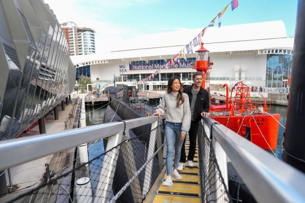 국립해양박물관 건물을 배경으로 붉은 배 근처의 다리를 걷고 있는 남성과 여성.