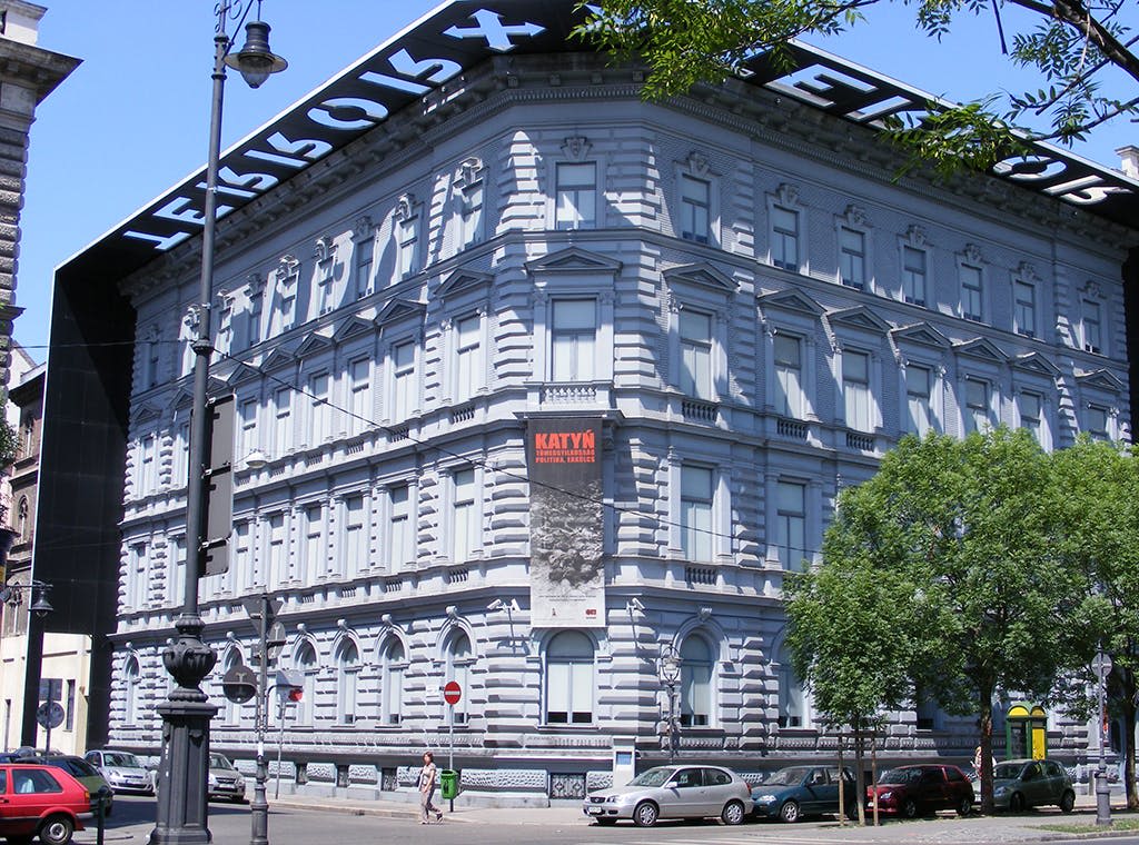 외관에 "KATYN" 이라고 적힌 표지판이 있는 크고 화려한 건물로 보행자와 주차된 차량이 있는 도시 거리에 있습니다.