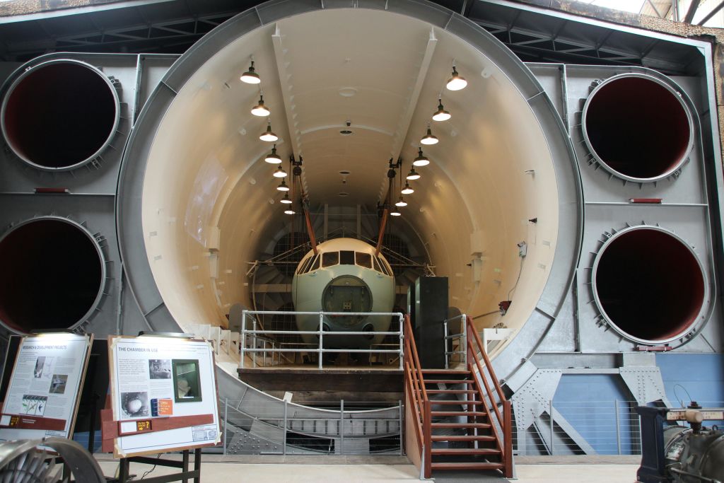 작은 항공기가 커다란 원통형 테스트 시설 내부에 배치되어 있으며 계단을 통해 올라갑니다. 오른쪽에는 두 개의 대형 배기 통풍구가 있습니다.