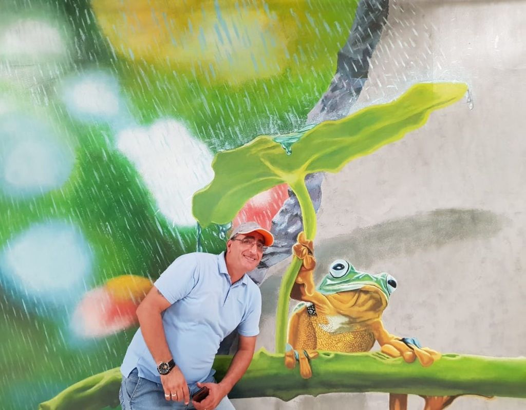 파란 셔츠를 입은 남성이 빗속에서 잎사귀를 들고 있는 개구리 벽화 옆에서 포즈를 취하고 있습니다. 밝고 화려한 배경.