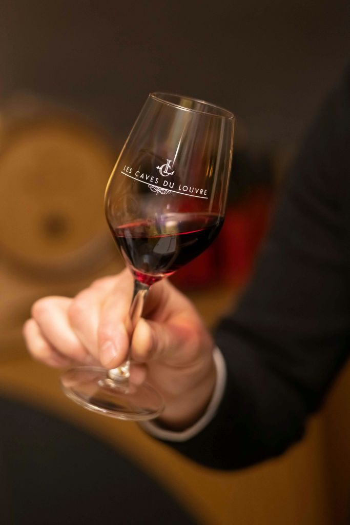 배경이 흐릿한 '루브르 동굴 (Les Caves du Louvre)' 이라고 적힌 레드 와인이 담긴 와인잔을 들고 있는 손.