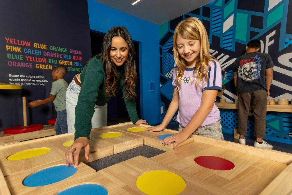 미소 짓는 어린 소녀와 어른이 다채로운 원이 있는 나무 탁자와 상호 작용합니다. 또 다른 사람이 배경에 있습니다.