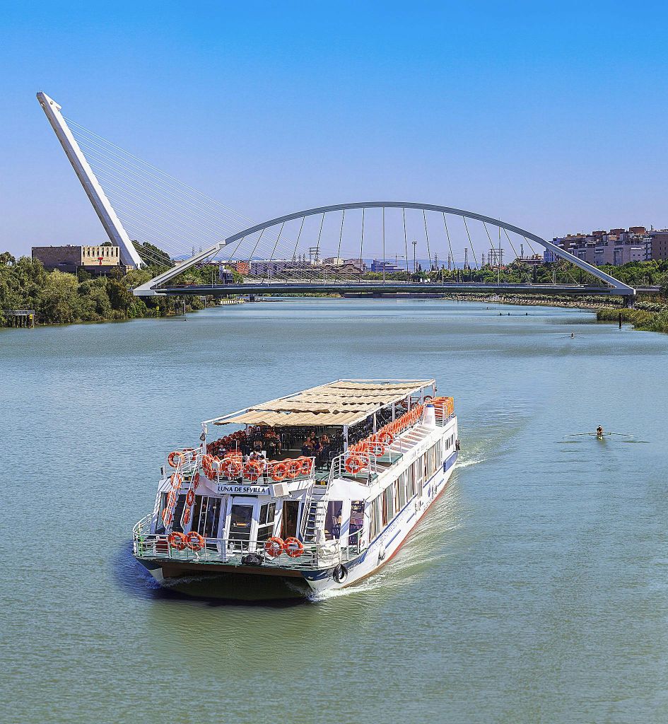 하얀 강선이 도시 건물과 푸른 하늘을 배경으로 케이블이 달린 현대적인 아치형 다리 아래를 항해합니다.