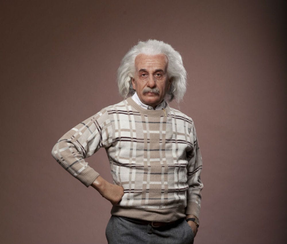 흰색 머리카락과 콧수염을 가진 노인이 베이지색 격자무늬 스웨터와 회색 바지를 입고 평범한 배경에 서 있습니다.