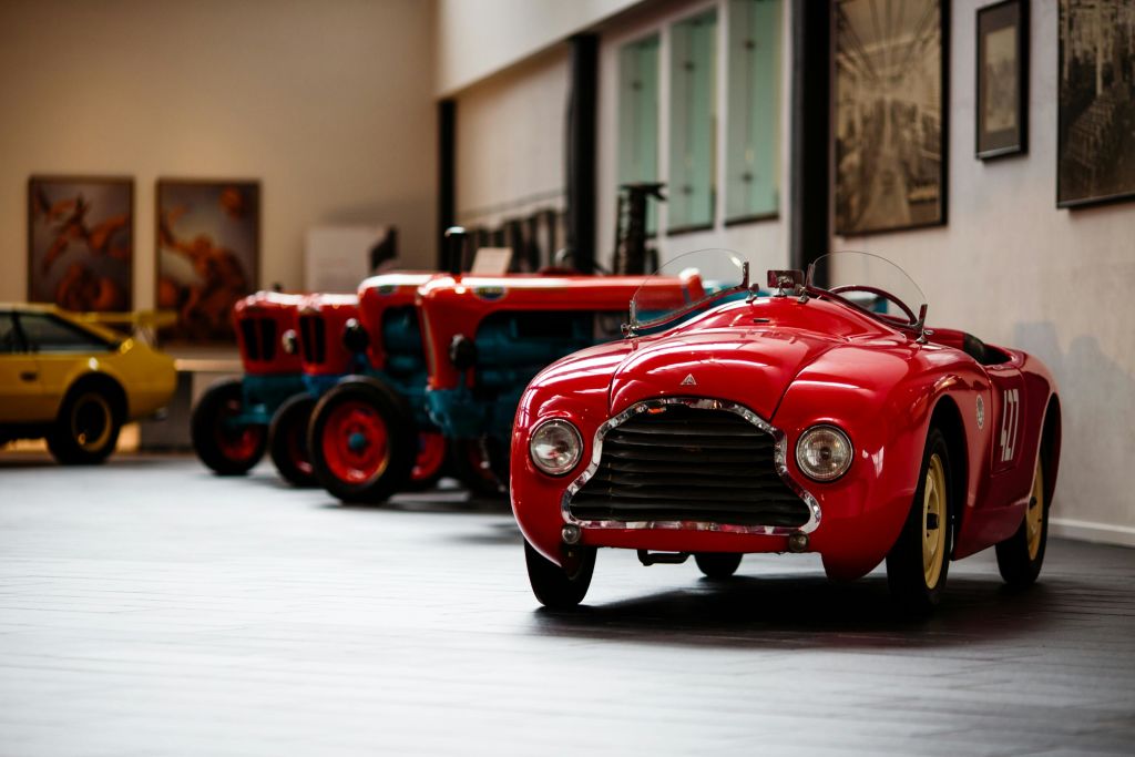 클래식한 빨간색 자동차가 박물관에 전시되어 있으며, 여러 대의 빈티지 파란색과 빨간색 트랙터가 배경에 줄지어 있습니다.