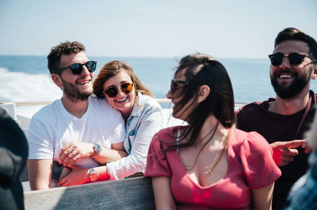 세 사람이 보트에 앉아 미소를 짓고 선글라스를 쓰고 있습니다. 바다와 푸른 하늘이 배경으로 보입니다.