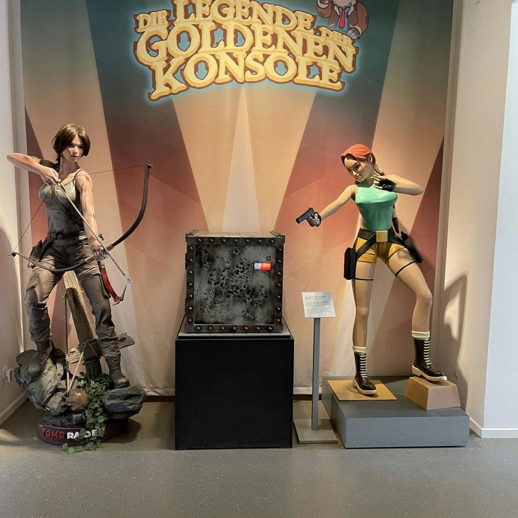 실물 크기의 여성 액션 피규어 조각상 두 개, 하나는 활과 화살, 다른 하나는 이중 권총이 금속 상자 옆에 전시되어 있습니다.