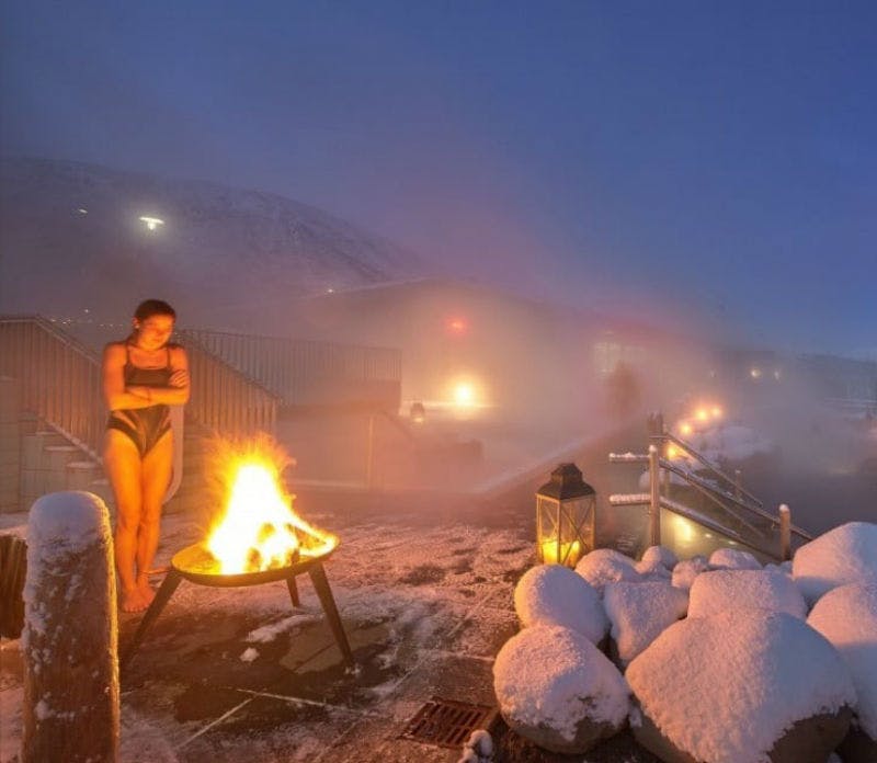 한 사람이 눈 내리는 날씨에 화덕 근처에 서서 수영복을 입고 팔짱을 끼고 안개와 불빛을 배경으로 서 있습니다.