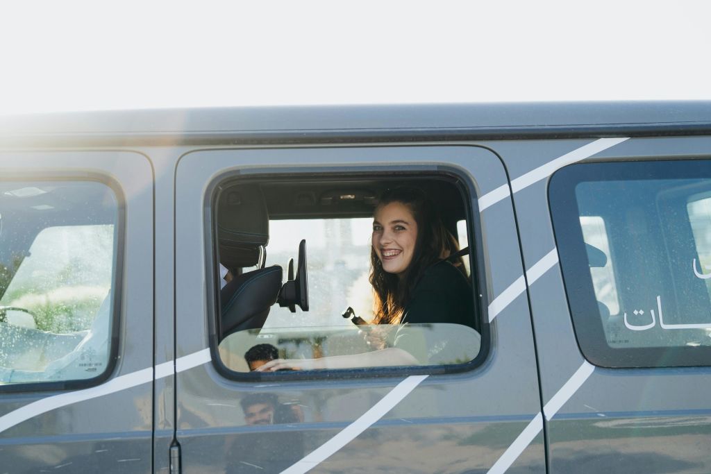 긴 머리를 한 사람이 열린 창문 너머로 보이는 차량 뒷좌석에 앉아 미소를 짓고 있습니다.