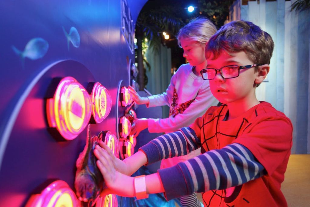 한 명은 줄무늬 셔츠를 입고 다른 한 명은 분홍색 셔츠를 입은 두 명의 어린이가 화려한 조명 버튼 벽과 상호 작용합니다.