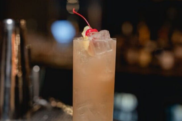 밝은 갈색 음료, 얼음, 체리, 레몬 웨지 장식으로 채워진 높은 유리잔이 희미한 배경에 맞춰져 있습니다.