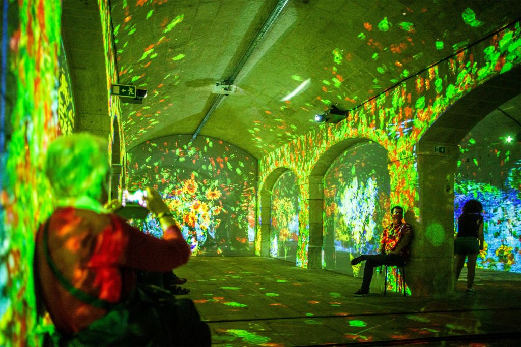 한 사람이 의자에 앉아 아치형 터널의 벽과 천장에서 다채로운 디지털 투영을 관찰하는 사람들.