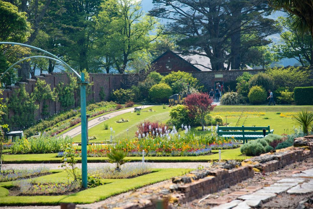 화려한 색상의 화단, 통로, 벤치, 나무가 있는 잘 관리된 정원으로, 건물 근처를 배경으로 사람들을 만날 수 있습니다.
