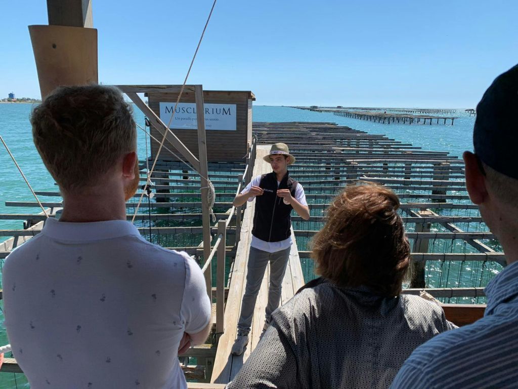 가이드가 나무 플랫폼에서 '무스클라리움' 이라고 적힌 표지판을 배경으로 두 사람에게 말을 건네고 있습니다.