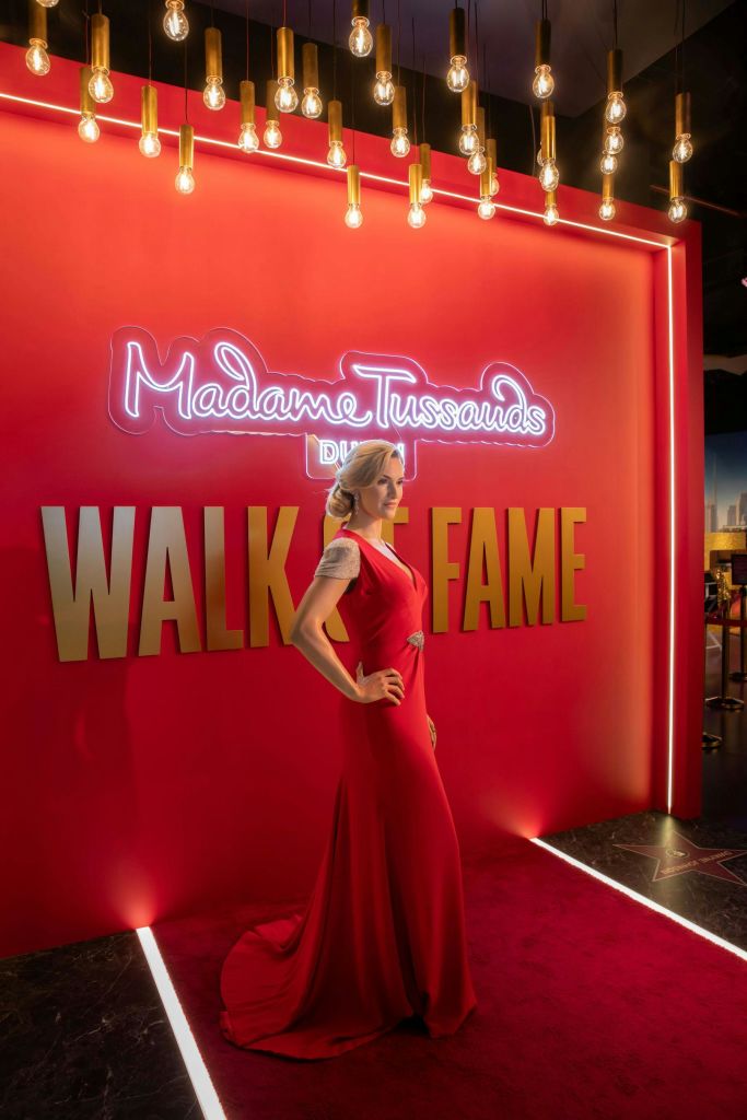 빨간색 드레스를 입은 여성이 네온사인 마담 투소 "명예의 전당" 표지판 앞에 서 있습니다.