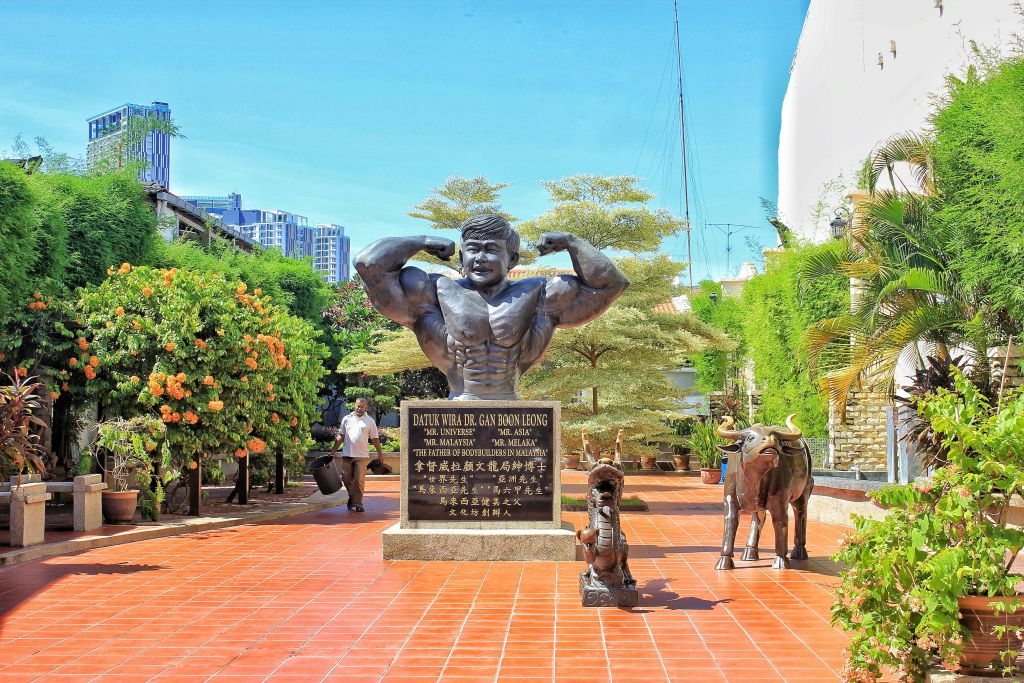 식물과 붉은 타일로 장식된 산책로에 둘러싸인 아래 판이 있는 근육질 남성의 동상. 인근의 작은 동물 조각상.