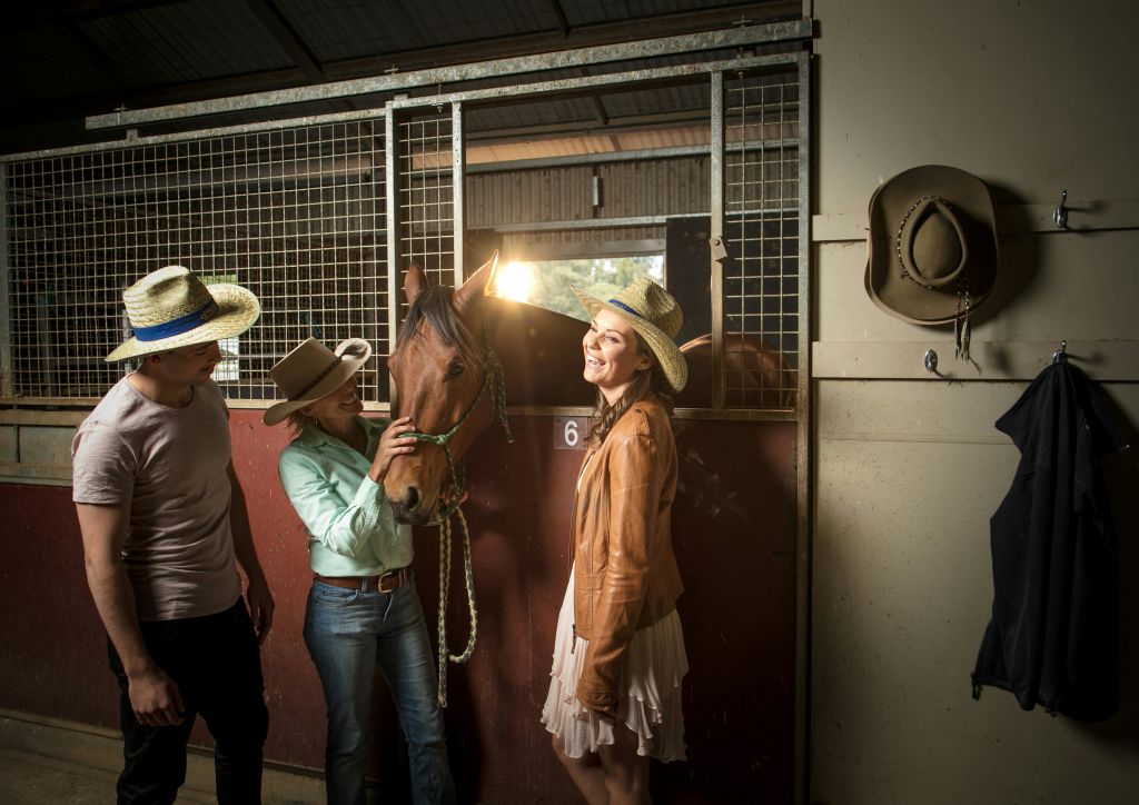 카우보이 모자를 쓴 세 사람이 마구간에 서서 갈색 말을 쓰다듬고 있습니다. 근처에 있는 벽에 모자 두 개가 걸려 있습니다.