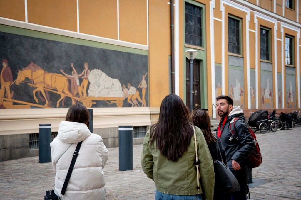 벽에 대형 벽화가 걸린 건물 앞에서 한 남성이 두 여성과 이야기를 나누고 있습니다. 근처에 자전거가 주차되어 있습니다.