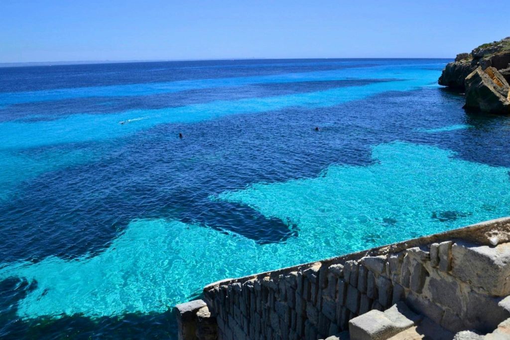 다양한 음영의 맑고 푸른 물과 전경의 돌담, 그리고 멀리서 수영하는 사람들이 몇 명 있는 해안 풍경.