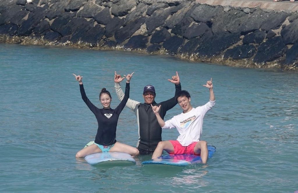 바위 해변 근처의 얕은 물에서 서핑 보드에 앉아 웃으며 손짓을 하는 세 사람.