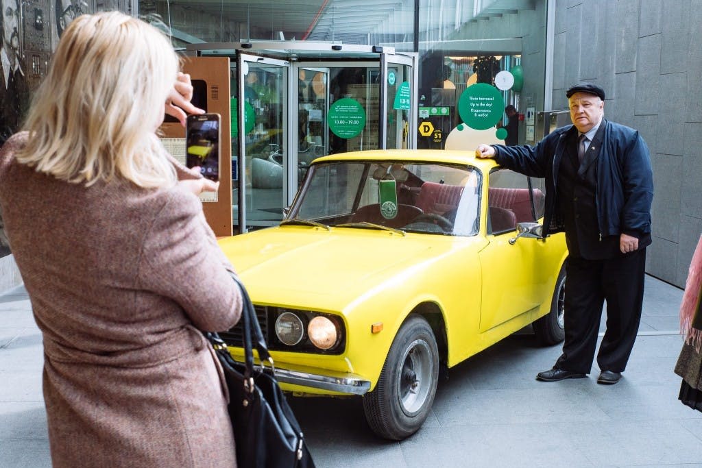건물 유리 입구 앞의 노란색 빈티지 차량 옆에 한 노인이 서 있습니다.