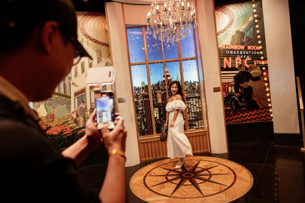 하얀 드레스를 입은 한 여성이 건물 내부의 도시 풍경을 배경으로 샹들리에가 머리 위로 놓인 사진 포즈를 취하고 있습니다.