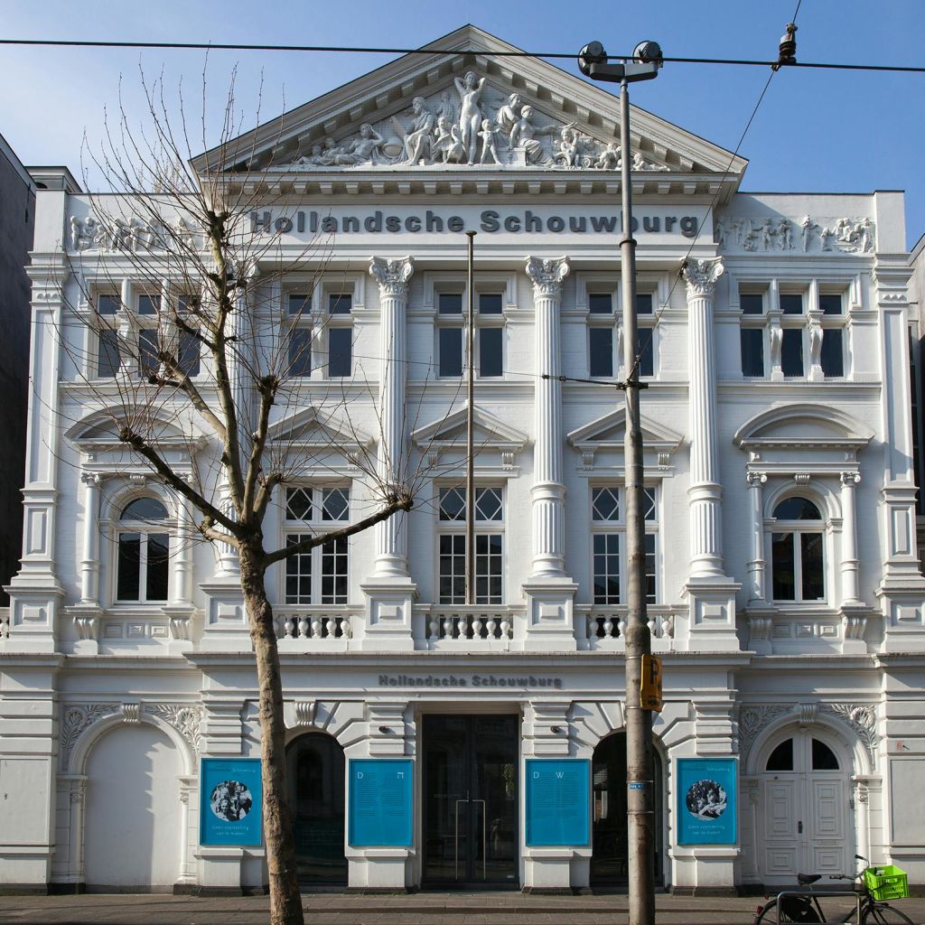 높은 기둥과 화려한 디테일이 돋보이는 유서 깊은 흰색 건물. 표지판에는 "Hollandsche Schouwburg" 라고 쓰여 있습니다. 앞에 잎이 없는 나무와 트램 와이어가 있습니다.