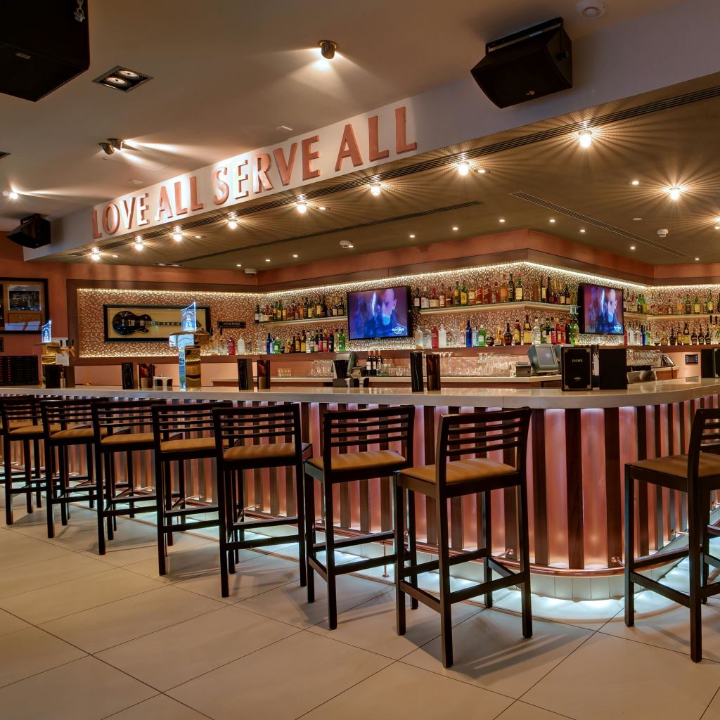 긴 카운터, 유아용 식탁 의자, 조명 선반, 두 대의 TV가 있는 바입니다. 위 표지판에는 'LOVE ALL, SERVE ALL.' 이라고
