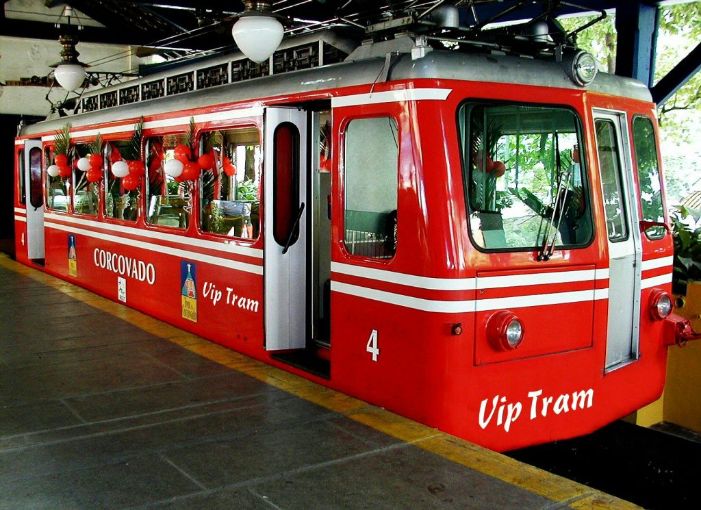 측면에 "Corcovado" 와 "Vip Tram" 이 적힌 빨간색 트램이 빨간색과 흰색 풍선으로 장식된 역에 주차되어 있습니다.