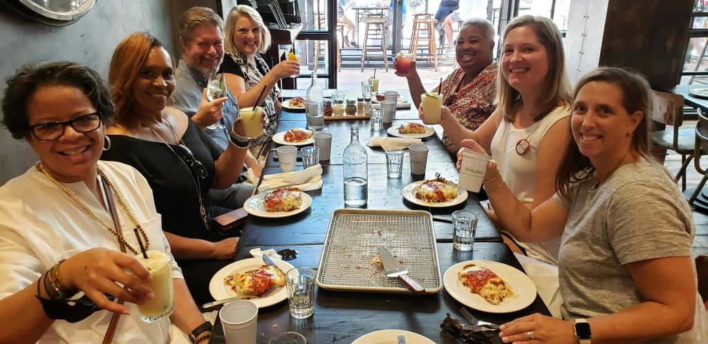 세 명의 여성이 레스토랑의 테이블에 앉아 음료를 들고 미소를 짓고 있으며, 테이블에 음식과 음료가 놓여 있습니다.