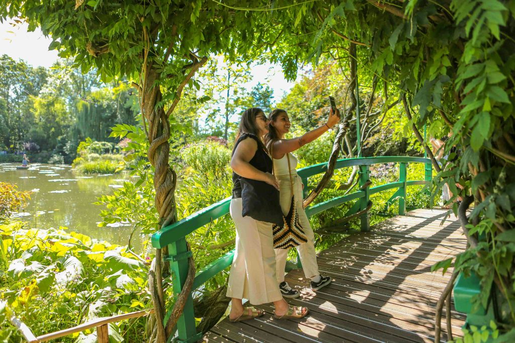 녹지와 덩굴로 둘러싸인 울창한 정원의 그늘진 나무 다리에서 두 여성이 셀카를 찍고 있습니다.