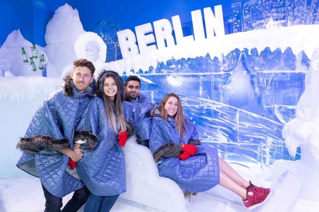 파란색 퀼트 코트를 입고 모피를 두른 네 명이 흰색 글씨로 된 '베를린' 이 있는 얼음을 테마로 한 방에서 미소를 짓고 있습니다.