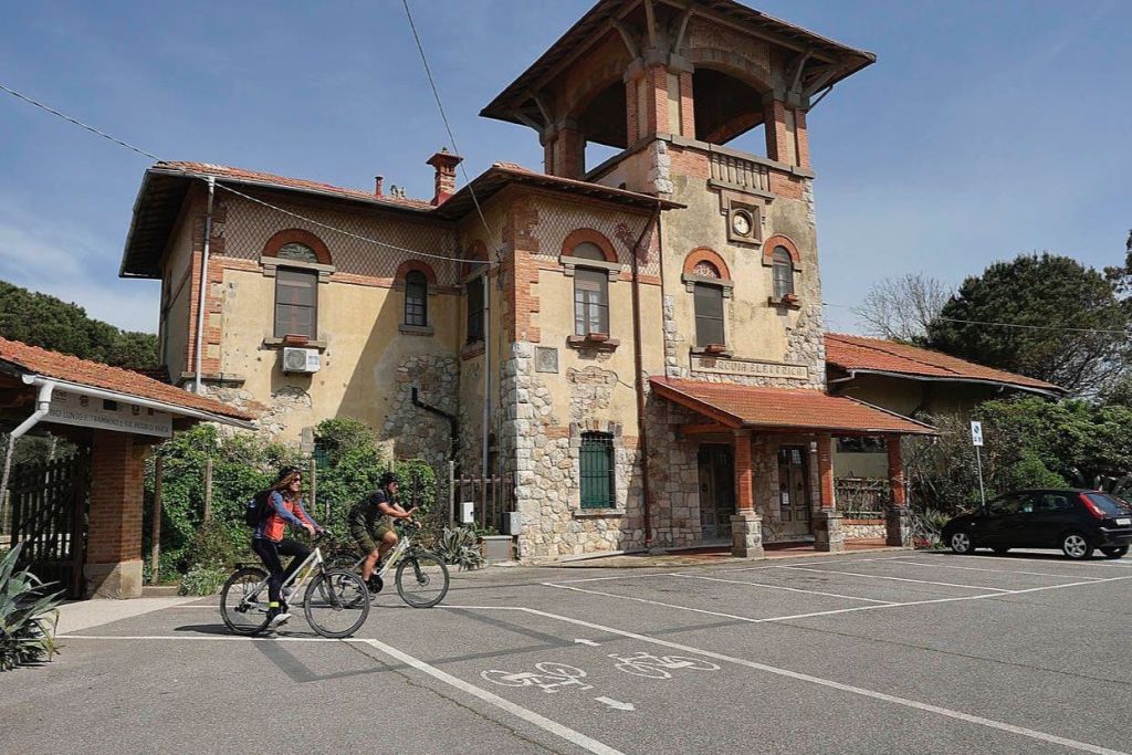 두 사람이 자전거를 타고 아치형 창문과 붉은 타일로 마감된 지붕이 있는 소박한 복층 건물을 지나갑니다.