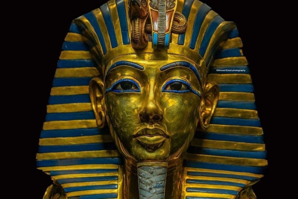 검은색 배경에 파란색과 금색 줄무늬 헤드피스와 복잡한 얼굴 디테일이 돋보이는 투탕카문의 황금 마스크.