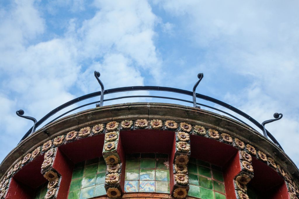 장식용 타일, 화려한 석공예품, 흐린 푸른 하늘을 배경으로 한 금속 난간이 있는 건물 외관의 상단 부분.