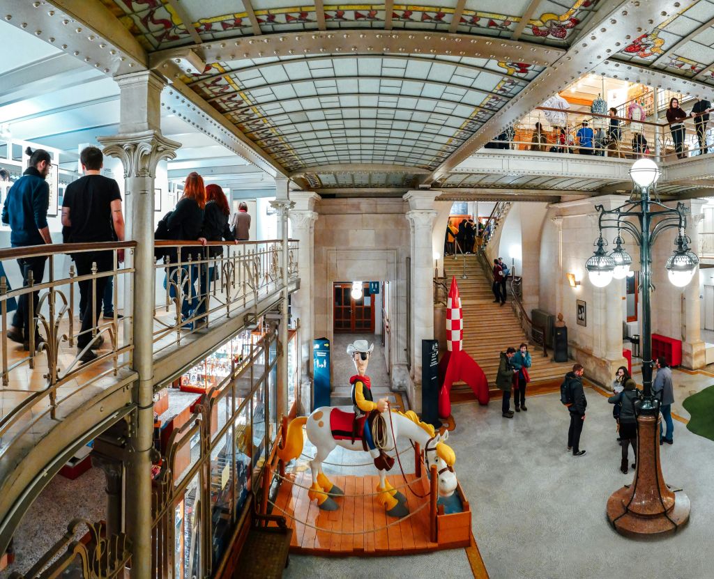 회전목마, 로켓선, 복잡한 천장과 난간이 있는 밝고 다층적인 공간에 있는 사람들.