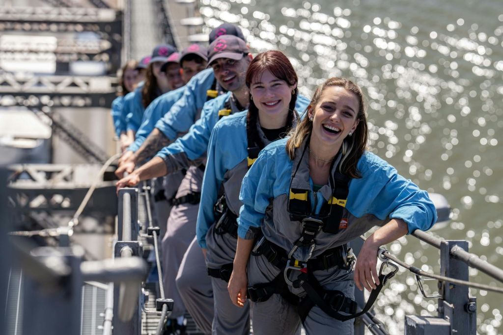 물을 배경으로 한 높은 통로에 줄을 서 있는 안전 장비를 착용하고 미소를 짓고 있는 사람들.