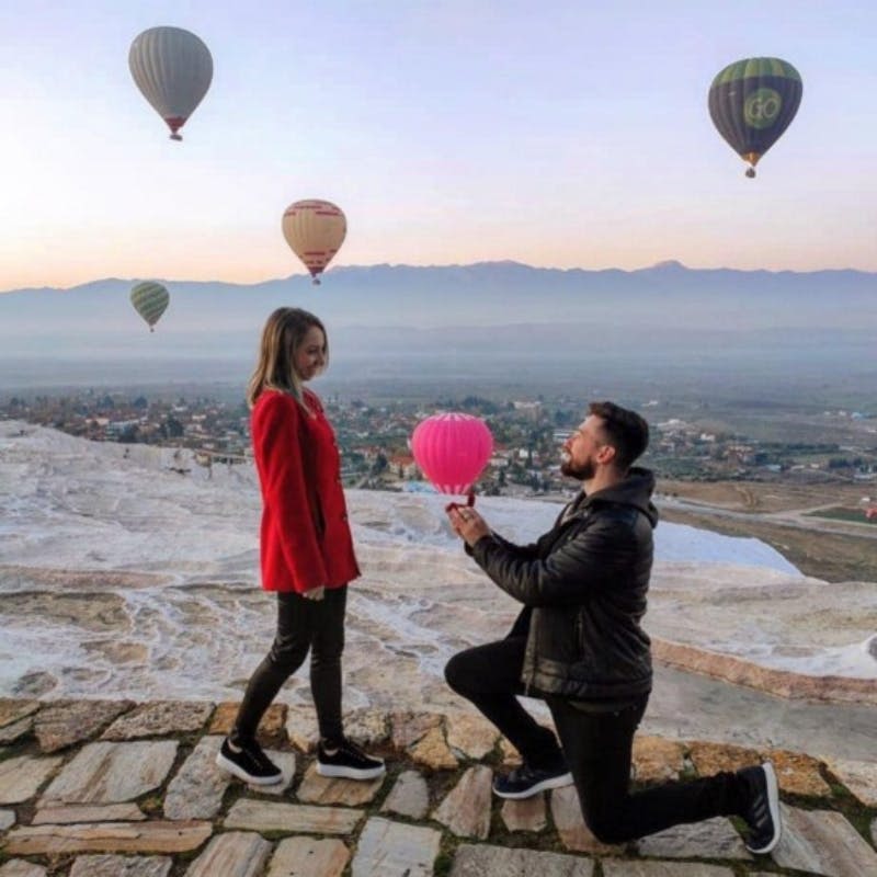 한 남성이 분홍색 풍선을 들고 하늘에 떠 있는 열기구와 함께 빨간색 코트를 입은 여성에게 제안합니다.