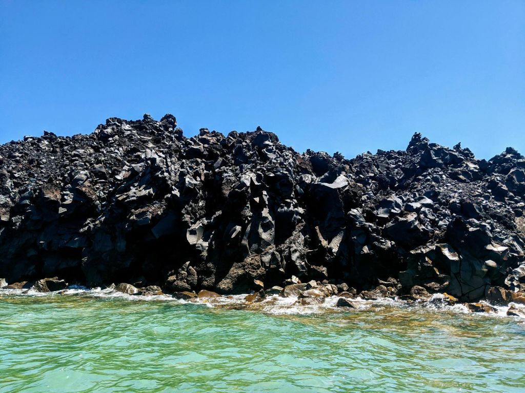 맑고 푸른 하늘을 배경으로 바위처럼 검은 용암층이 형성되어 있으며, 그 밑바닥에는 부드러운 바다 파도가 일렁이고 있습니다.