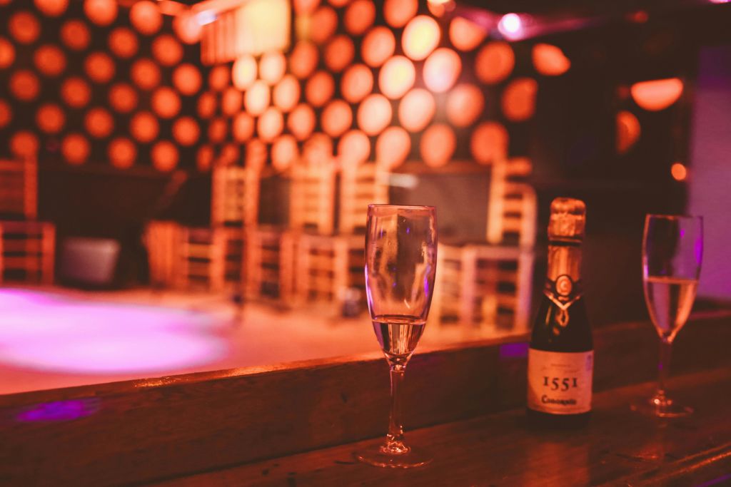 의자와 장식용 원형 패턴 벽이 있는 어두운 조명 방의 테이블에 샴페인 한 잔과 "1551" 이라는 라벨이 붙은 병.