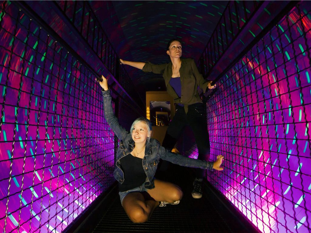 활기찬 보라색과 파란색 조명과 메쉬 측면이 있는 터널 안에 한 명은 웅크리고 서 있고 한 명은 서 있는 두 사람.