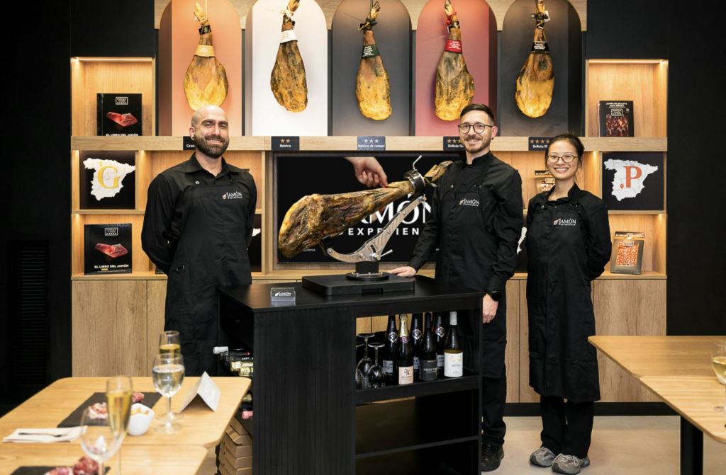 검은 유니폼을 입은 두 사람이 경화 햄, 디스플레이 카운터, 음료를 매달아 놓은 세련된 인테리어에 서 있습니다.