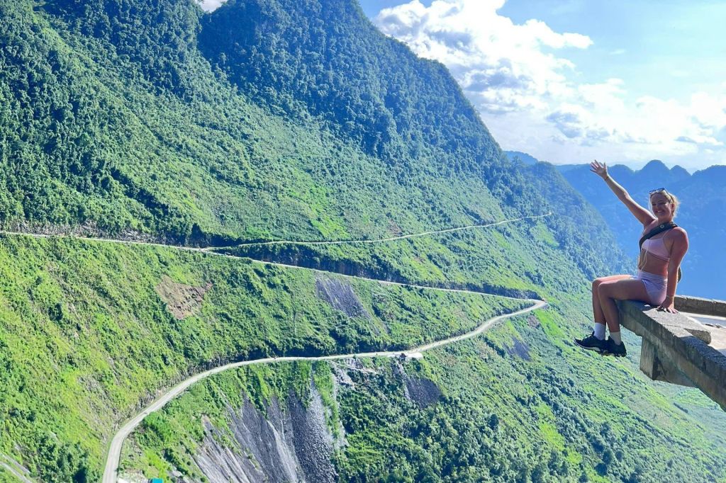울창하고 푸른 산비탈과 밝은 하늘 아래 구불구불한 도로가 한눈에 내려다보이는 바위 가장자리에 앉아 있는 사람.