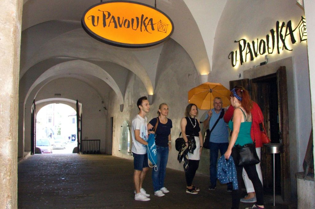 한 무리의 사람들이 "u Pavouka" 라고 적힌 간판이 있는 아치형 통로 아래에 서 있습니다. 한 사람이 노란 우산을 들고 있습니다.