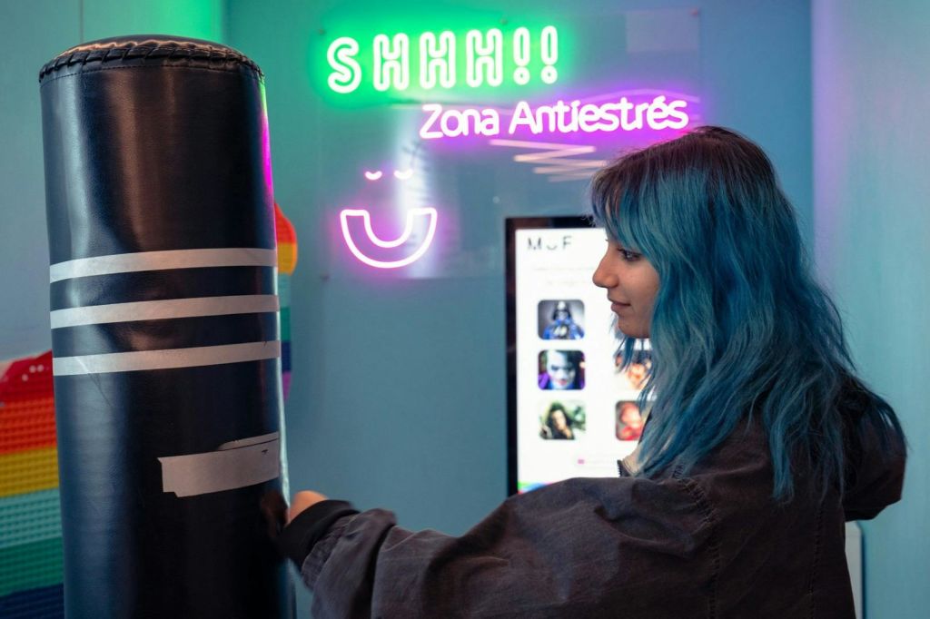 파란색 머리카락을 가진 사람은 화려한 방에서 "쉿!!" 과 "Zona Antiestrés" 를 읽는 네온 사인 근처에 서 있습니다.