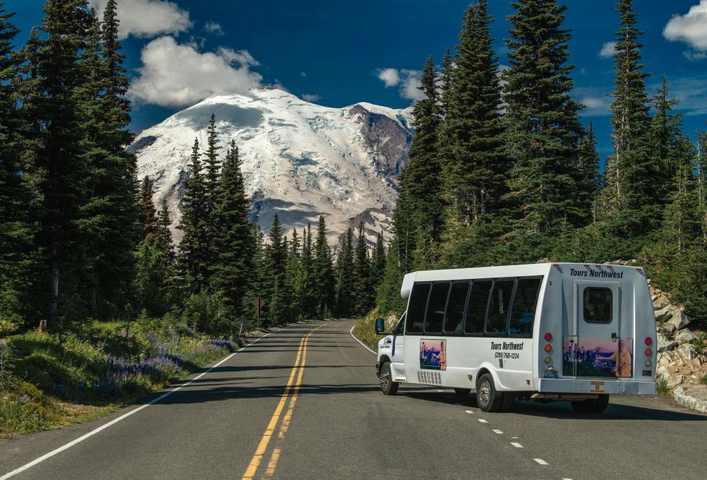 저 멀리 푸른 하늘 아래 눈 덮인 산과 함께 나무와 야생화로 둘러싸인 도로 위를 달리는 투어 버스.
