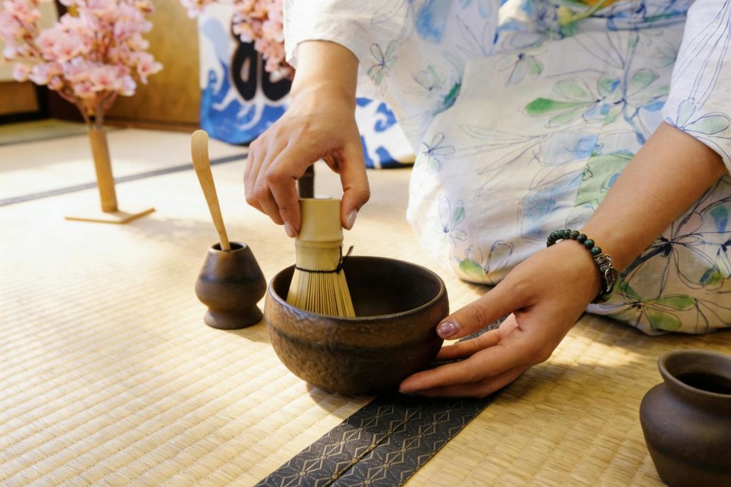 꽃무늬 인테리어가 돋보이는 밝은 방에서 전통 매트를 깔고 수염, 그릇, 숟가락으로 말차를 준비하는 사람.