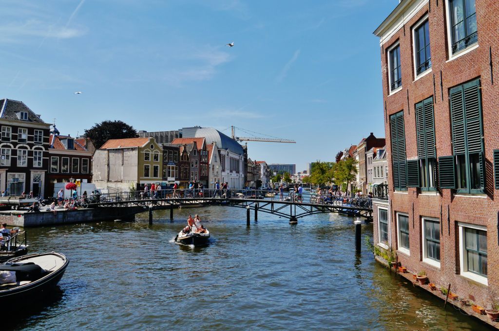 보트가 있는 운하, 보행자가 있는 다리, 역사적인 건물, 맑은 하늘 아래 크레인이 배경으로 있습니다.