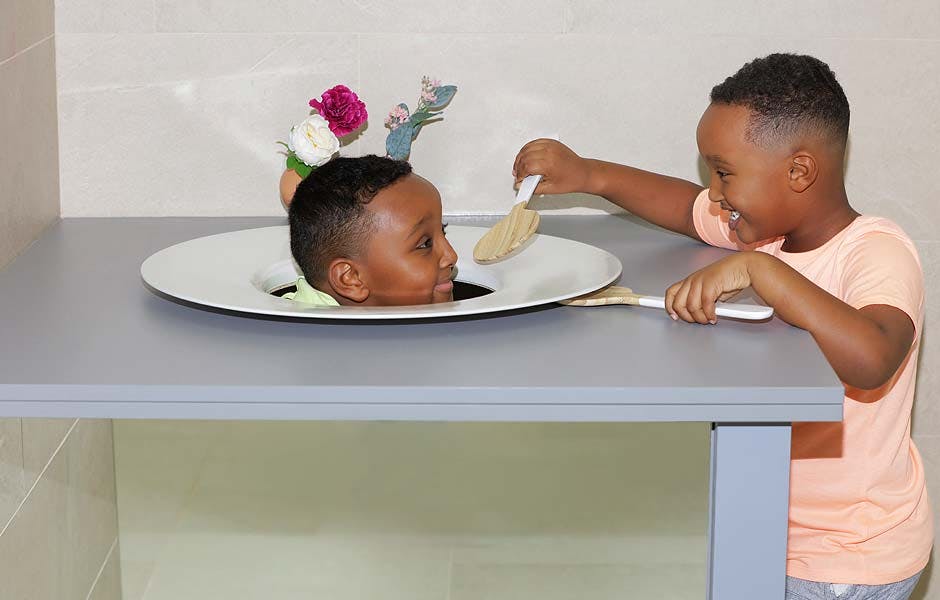 접시에 머리를 얹은 소년이 테이블을 통해 나타나고, 다른 소년은 장난스럽게 그에게 식기와 함께 음식을 제공합니다.